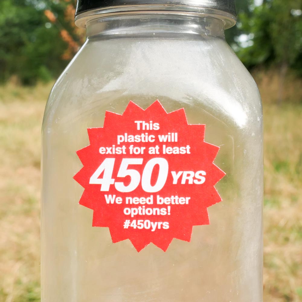 Photograph of a single-use plastic bottle with a red, starburst-shaped 1.25 inch sticker on it that says: This plastic will exist for at least 450 years. We need better options #450yrs. In the background, behind the bottle, is a green field