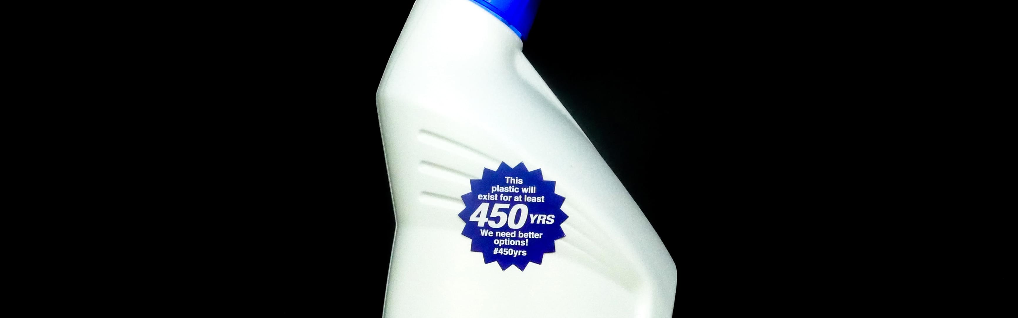 Close-up photograph of a blue plastic bottle, shaped like a liquid soap container. On the front of the bottle is a magenta, sunburst-shaped sticker that says, 'This plastic will exist for at least 450 YRS. We need better options! #450yrs.'