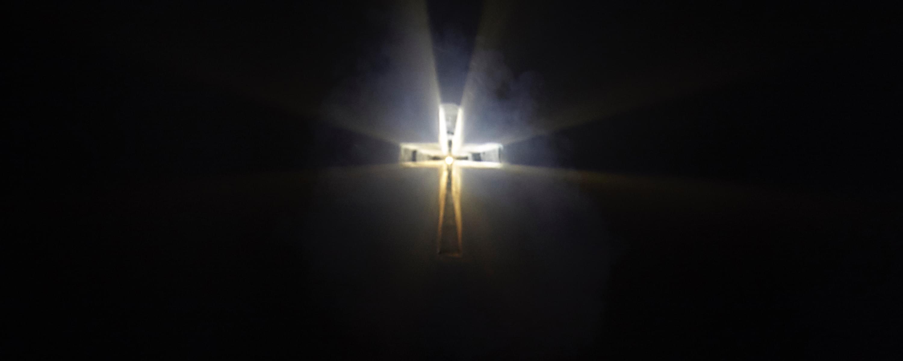 Photograph of a crucifix, hovering in mid-air and being formed soley by light. It is cloudy around the edges and there is a sharp circular focus of light in the middle from whence the light is shone.