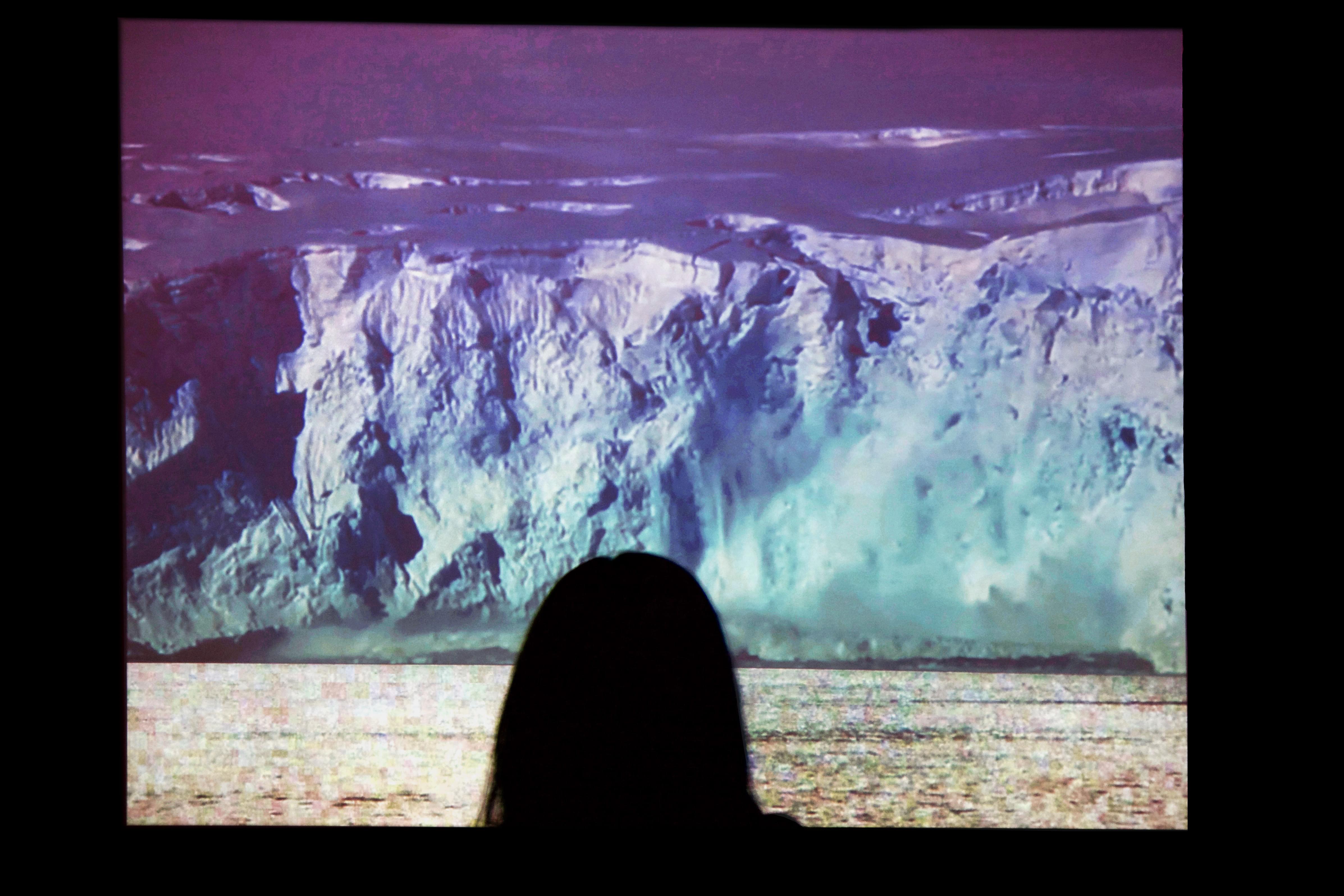 Close-up photograph of the back of a gallery-goer's head as they look at a projected image of a large glacier calving in front of them