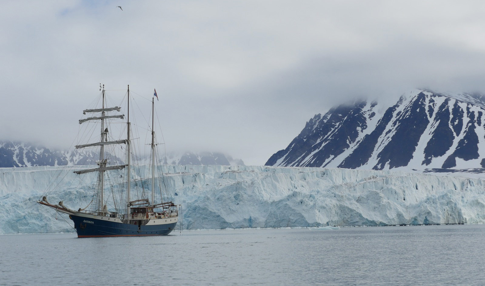 Photograph of an arctic landscape: cold gray sky, snow covered mountains in the far background, a 20-30 meter high glacier in the near-background, and a old-timey sail ship anchored in calm waters on the left of the frame.
