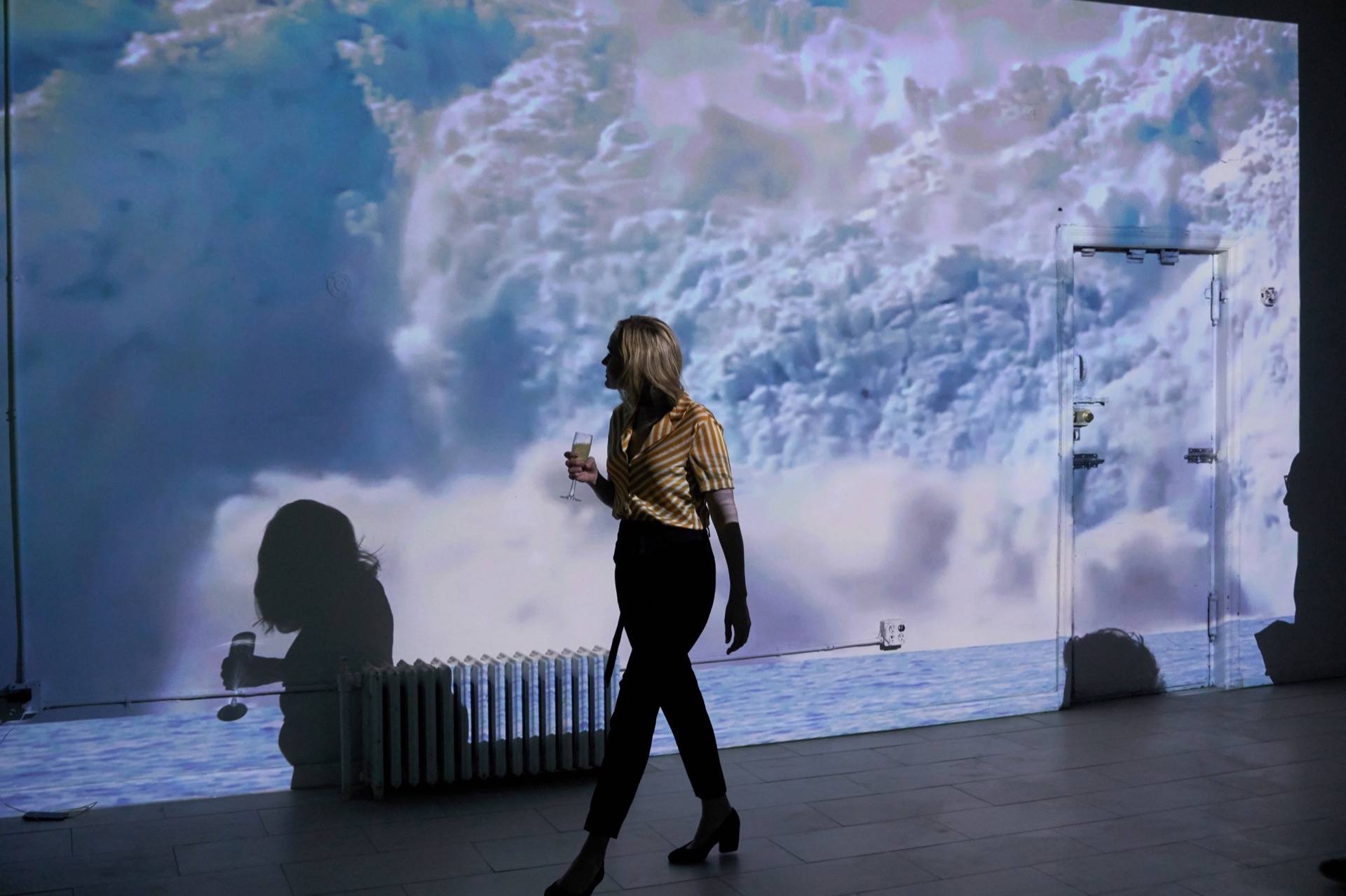 Small thumbnail image of Slighly askew photograph of a women in cocktail attire, holding a cocktail, walking in front of a projection of a glacier collapsing
