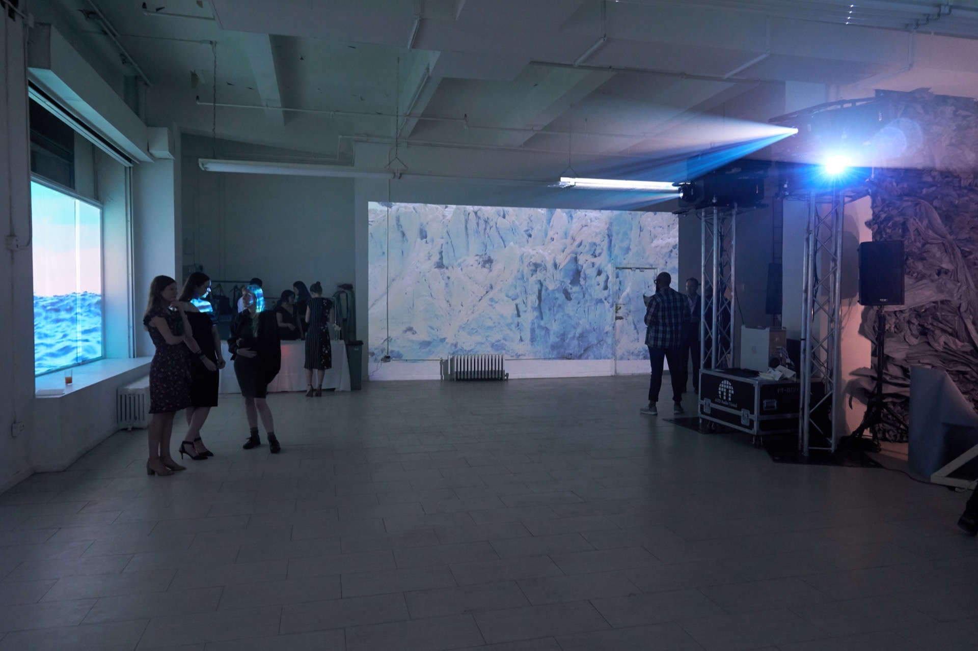 Small thumbnail image of Wide shot of the inside of a gallery showing two different projected works. On the left, an image of a body of water is projected into the gallery windows. Straight back, an image of an Arctic scene—close up of a glacier—is projected. There are about a dozen people in cocktail attire milling about. On the right, a two sharply bright lights are emanating from the lens of large projectors.