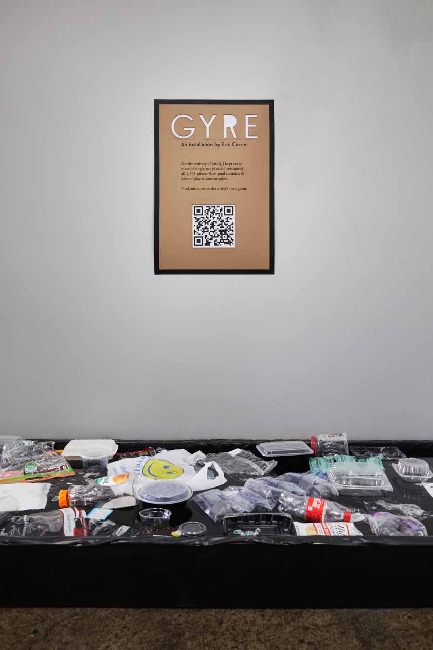 On a gray wall hangs an 18 inch by 24 inch sign that says 'GYRE' followed by some text and a QR code underneath. On the floor, perpendicular to the frame is a one foot high black pool with dozens and dozens of single use plastic items floating on the surface.