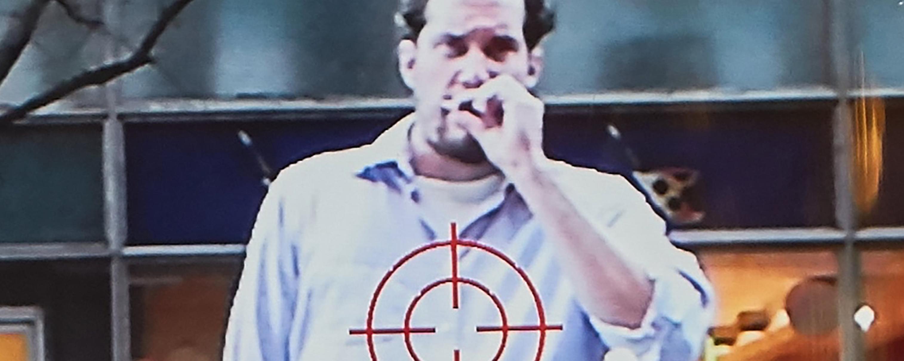Close up grainy photograph of a man smoking a cigarette in a white button down shirt, with a cross-hairs target symbol shown on top his shirt