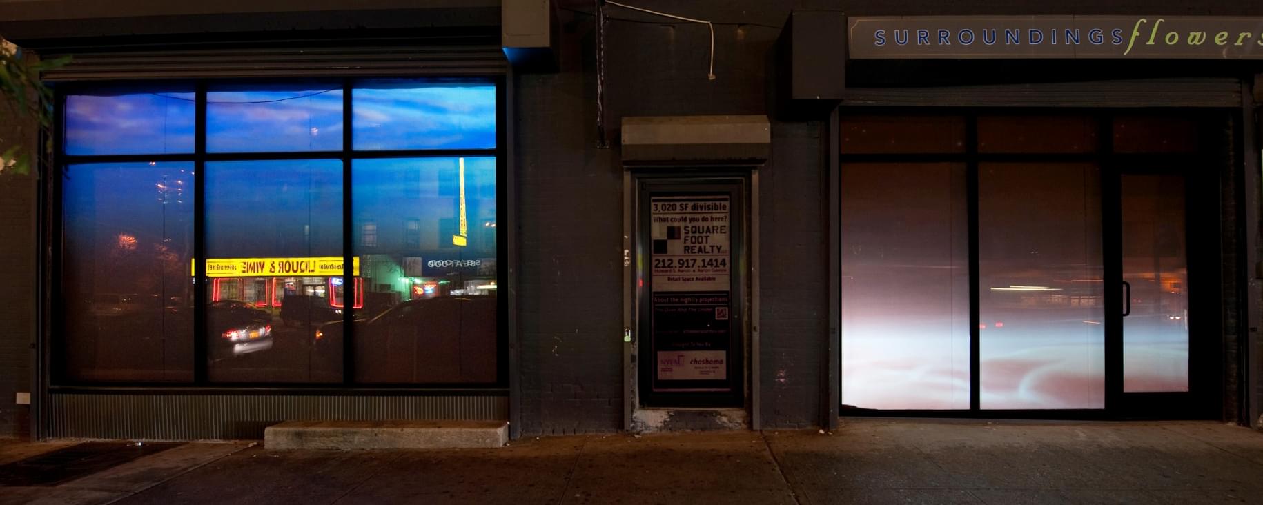 Nighttime photograph of two storefronts, one on each side of the frame, separated by an abandon-looking door in the middle. In the left storefront is projected an video of the surface of water, from below; reflected in the windows is a bright yellow awning of the liquor store across the street. In the right storefront is projected a violet-ish rendering of clouds as seen from above.