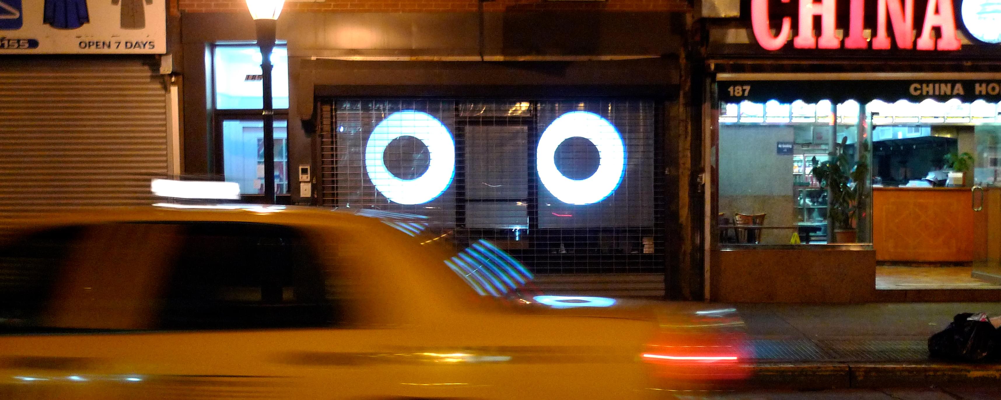 Nighttime photograph of a blurred NYC yellow cab moving across the frame with the two "Watcher" eyes in the storefront behind following it as it passes by
