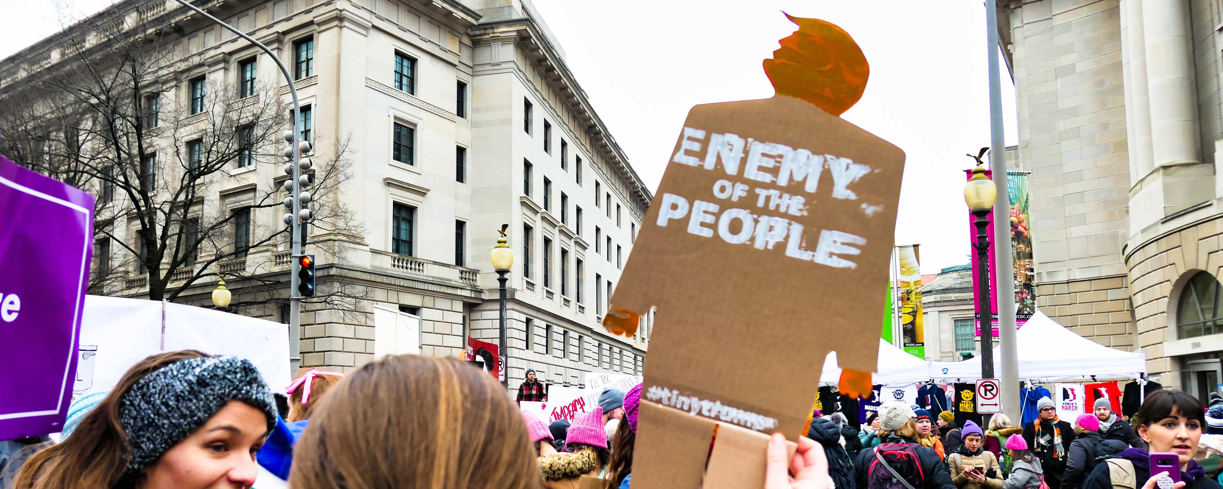 Photograph at a protest march of a women holding a tiny trump (2 foot tall cardboard cutout of trump) that has 'Enemy of the People' stamped on it in white ink