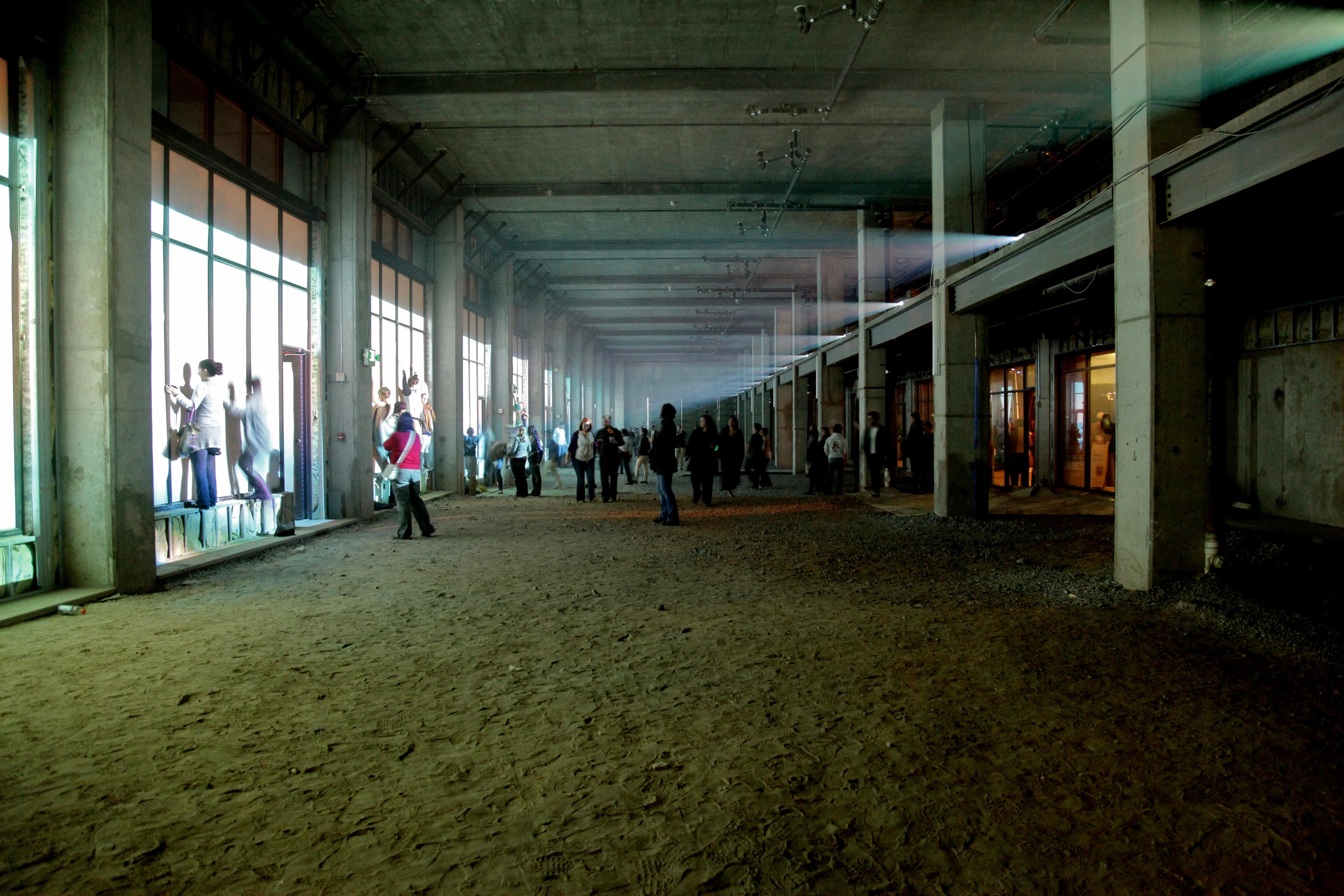 Photograph of a relatively dark, cavernous space (like the inside of an large, empty commercial building) with art festival goers milling about, some of whom have climbed into the windows to interact with what's being projected onto them (the water scene described above). On the upper left of the photograph one can see a line of about a dozen projectors shining light across the empty space onto the windows.