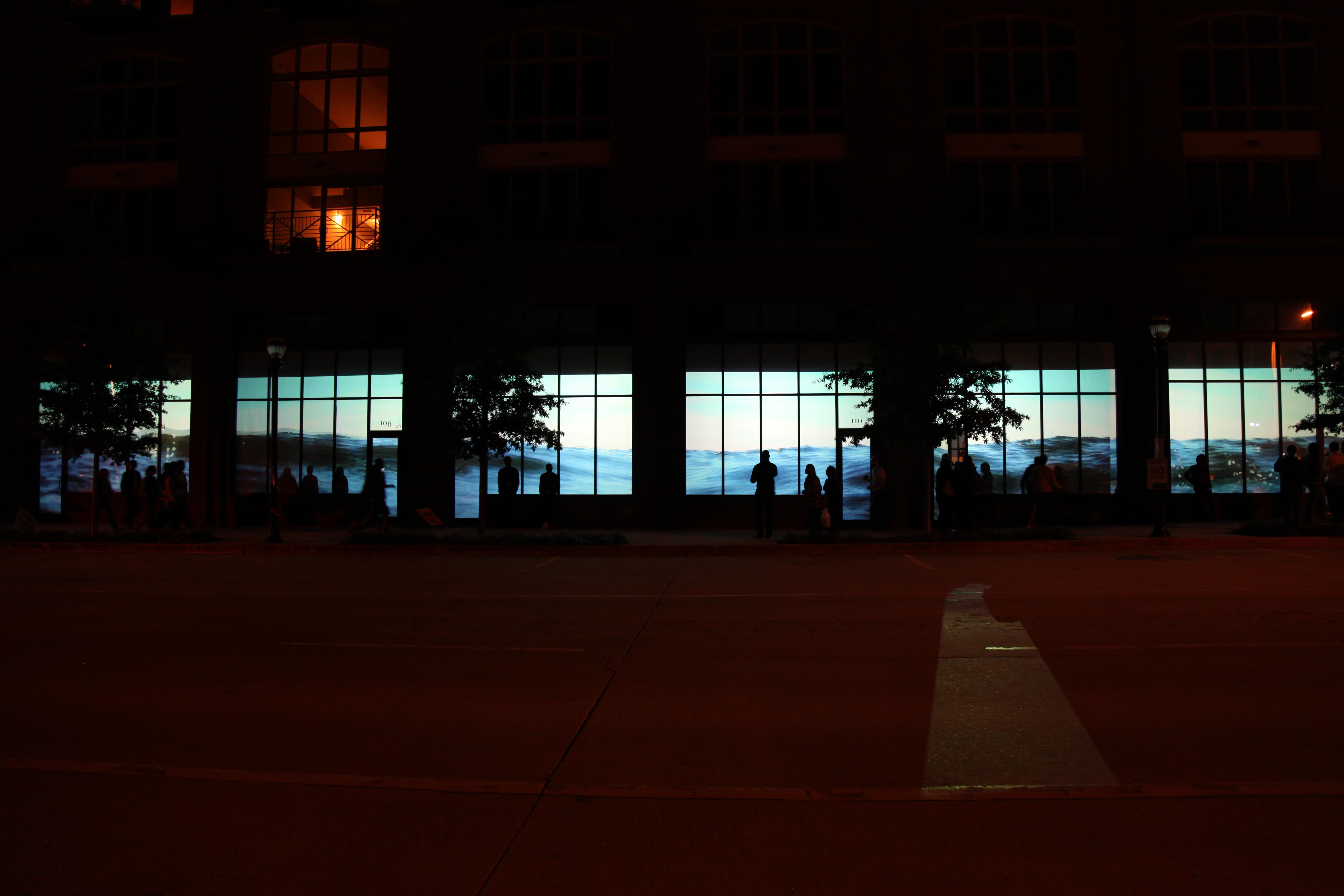 Small thumbnail image of Nighttime photograph of a large commercial center with six of its street-level storefront windows showing a contiguous video projection of water ebbing and flowing.
