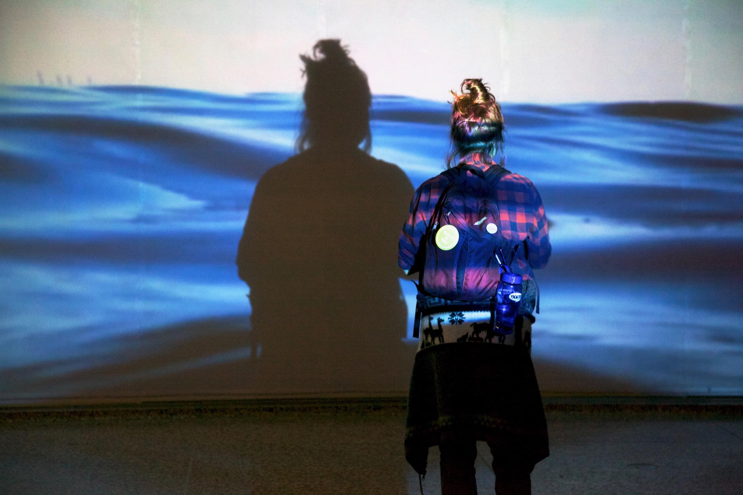 Small thumbnail image of Photograph of the back of a seemingly teenage girl wearing a backpack staring at the life-size projection of water in front of her. We see her shadow in the projection of the water.