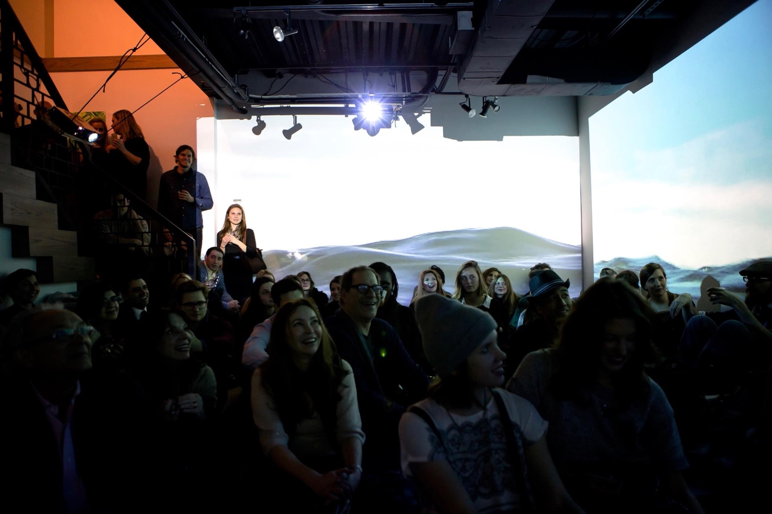 Photograph of a crowd of people sitting down in a dark gallery space with a body of water projected on the walls behind