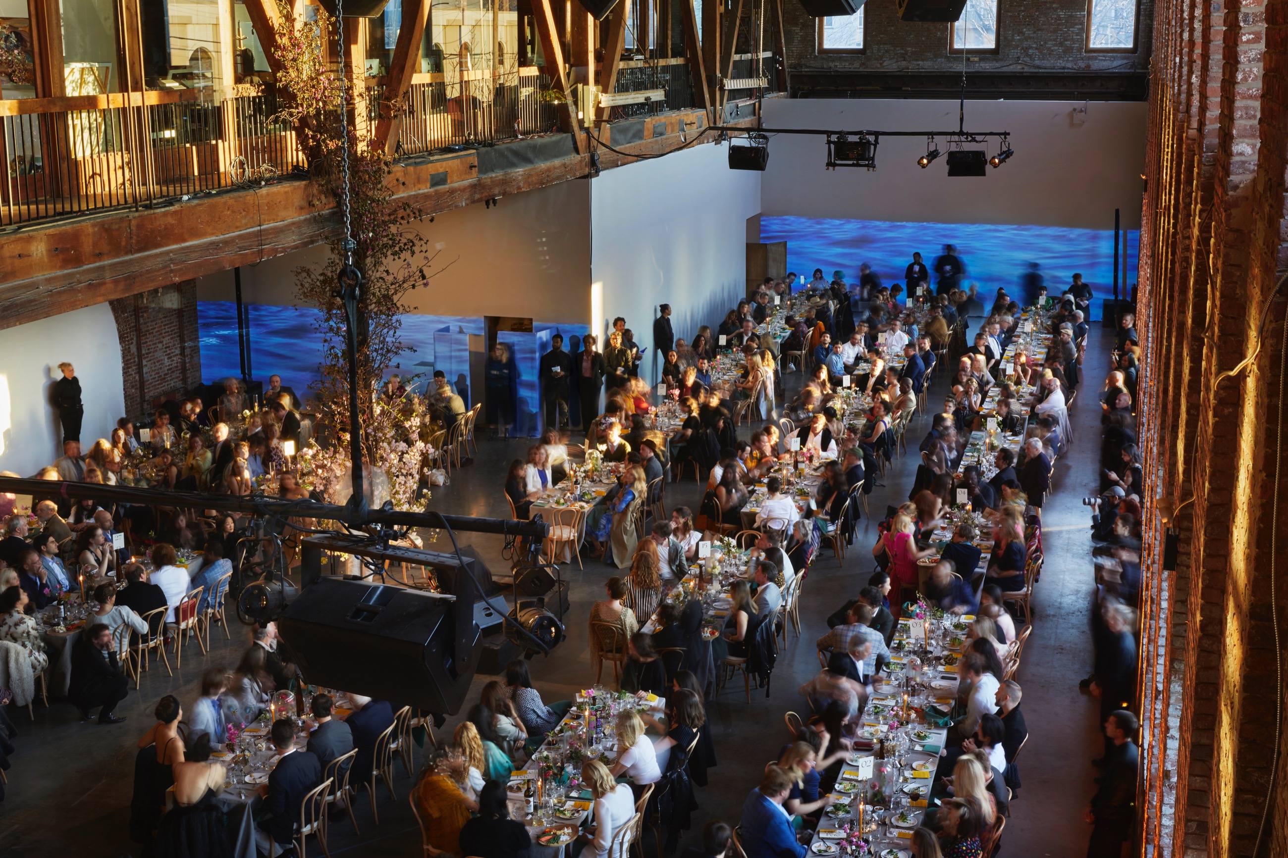 Overhead shot of a gala event—dozens of elegently decorated tables with white table cloths and well-dressed people sitting at them. Behind them, on a long white wall, a video of a body of water is projected