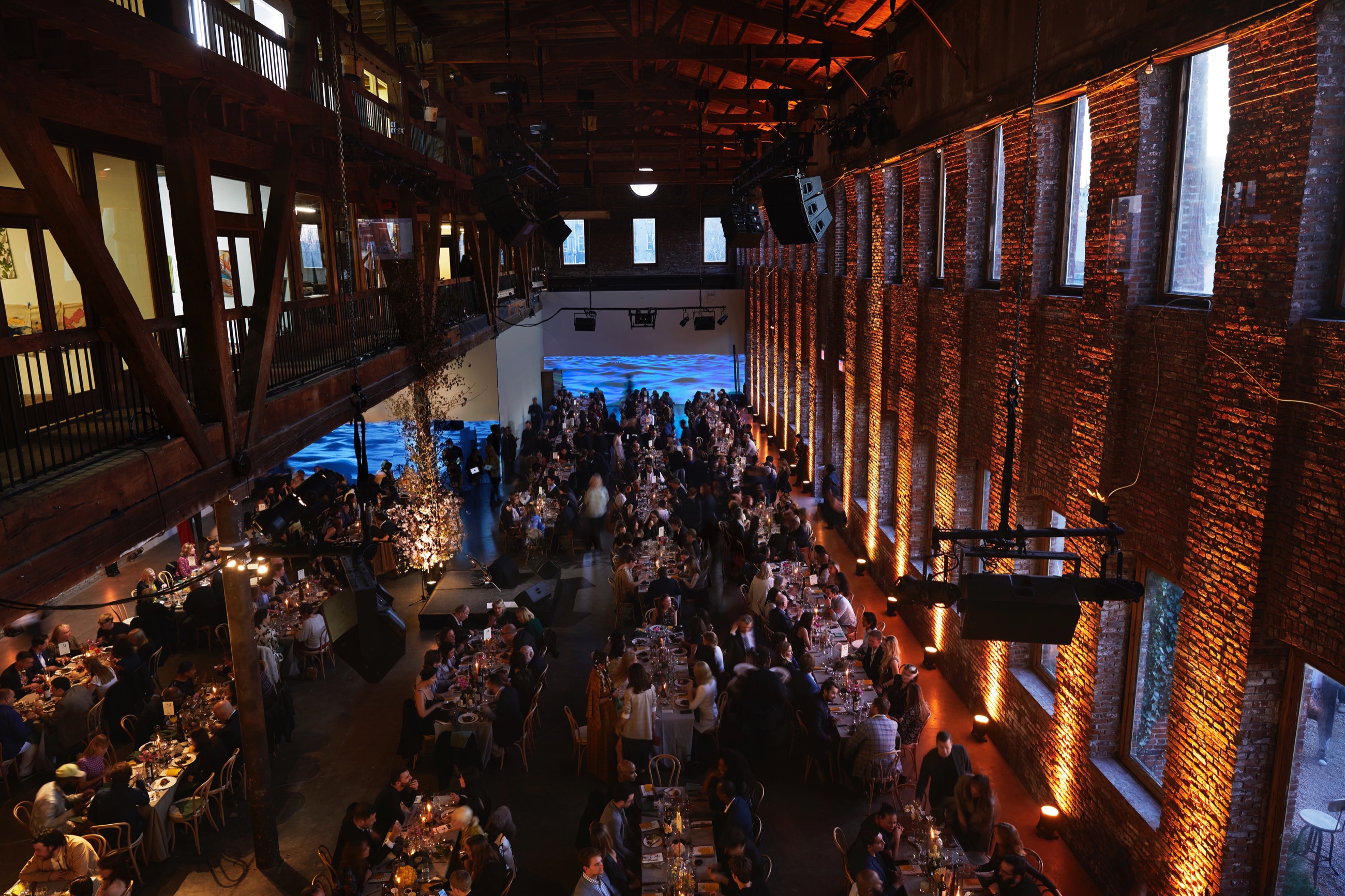 Overhead shot of a gala event, over elegently decorated tables with well-dressed people seated at them. Behind them, on a long white wall, a video of a body of water is projected 