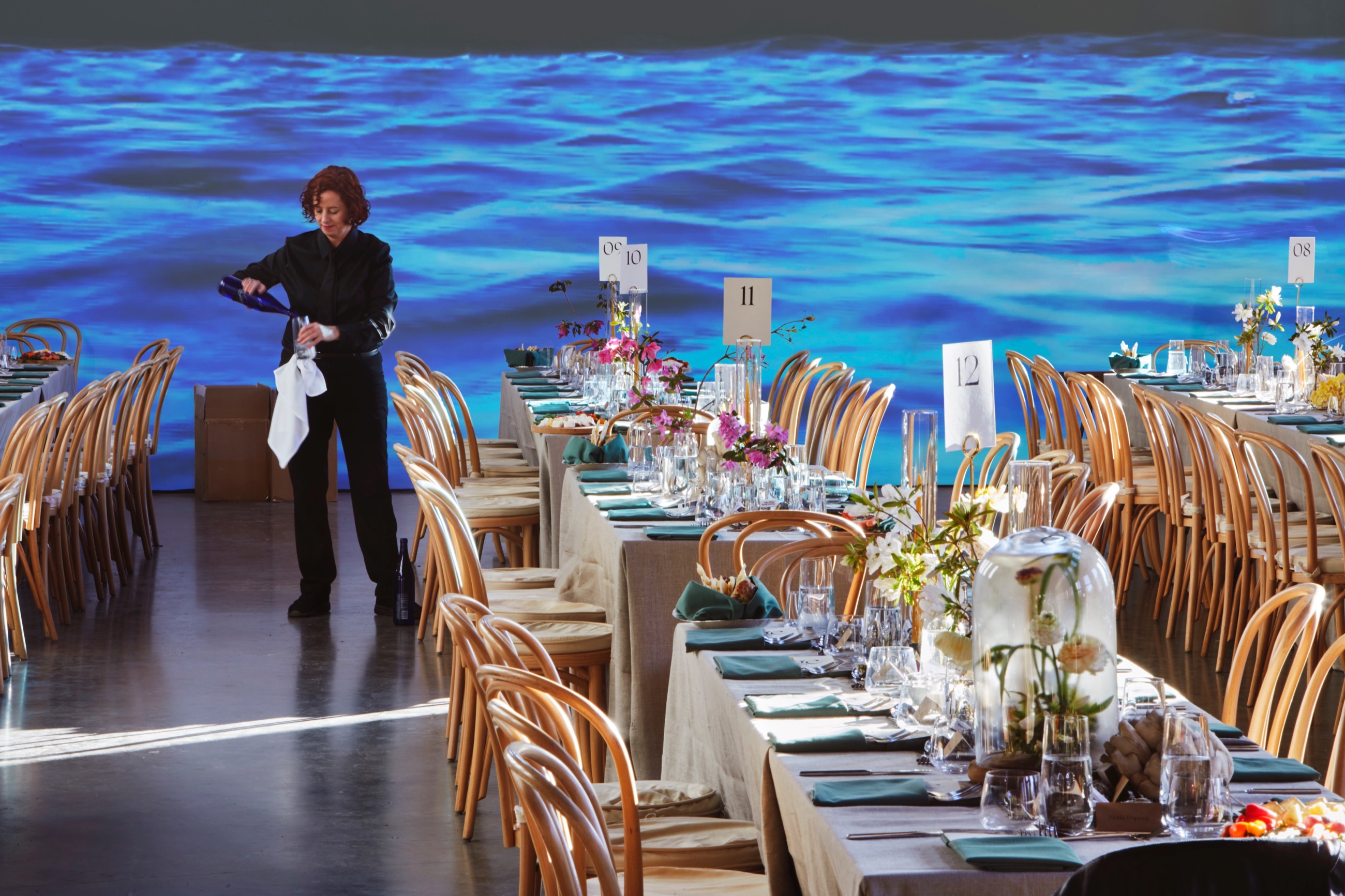 Small thumbnail image of Foreground: white table clothed tables with numbers suggesting a gala event, though no one is seated at them yet. Middleground: a server pours water into a glass. Background: a video of a body of water is projected on a white wall.
