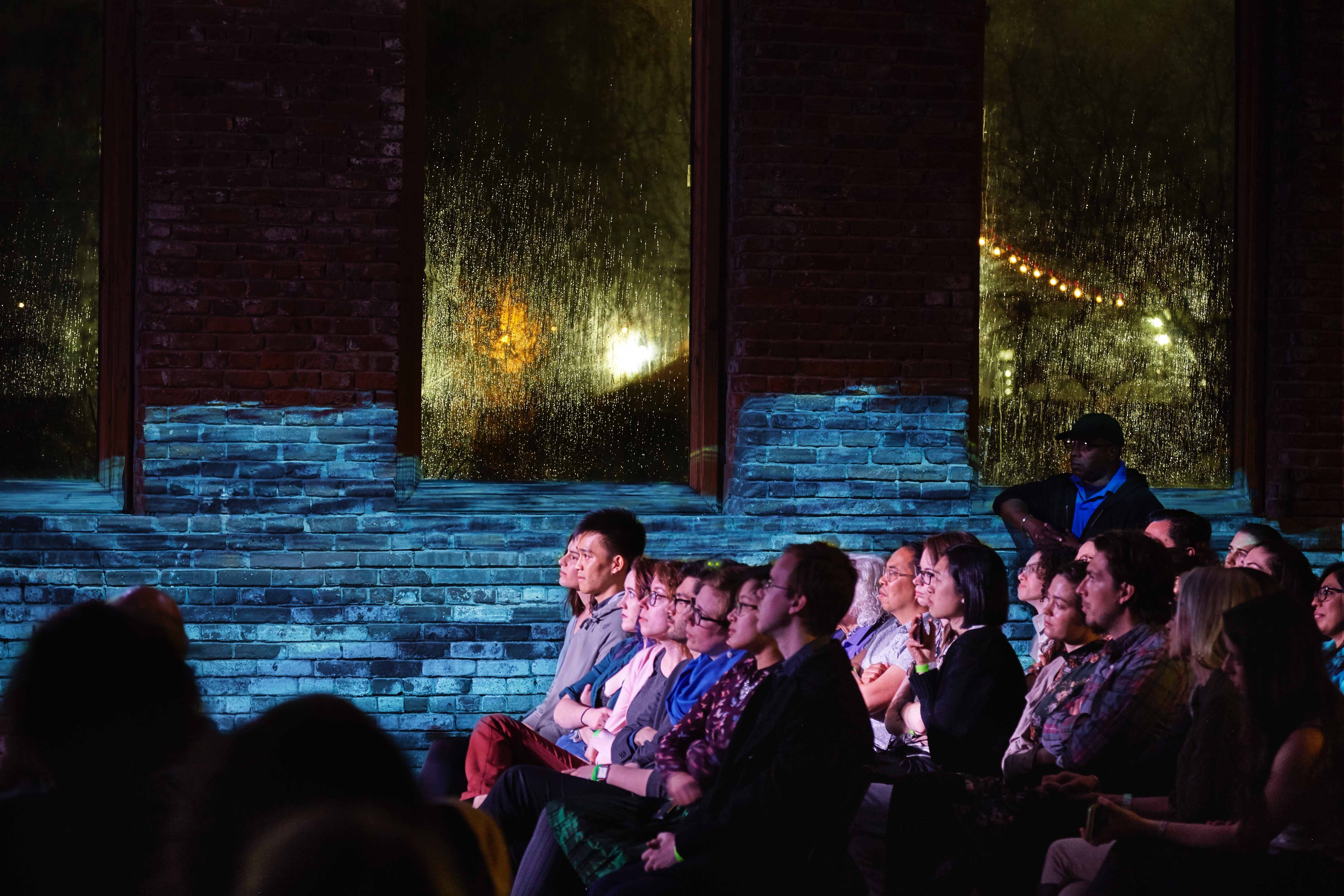 Photograph of three rows of audience shot in profile. A video of a body of water is projected on the brick wall behind them.