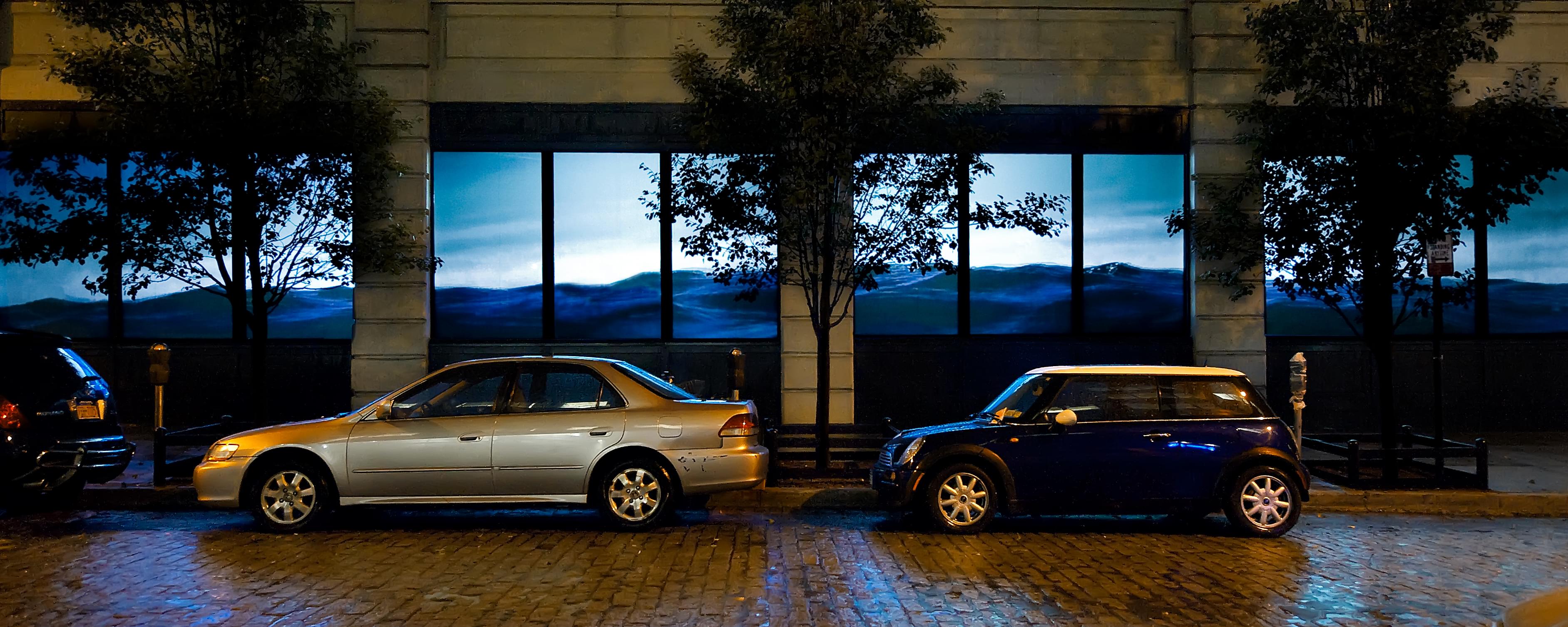 Photograph a building facade at night where, projected onto its street-level windows, is a eye-level view of small ocean waves, presumably bobbing up and down. In front of the building are two parked cars.