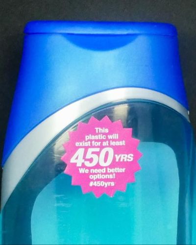 Close-up photograph of a blue plastic bottle, shaped like a liquid soap container. On the front of the bottle is a magenta, sunburst-shaped sticker that says, 'This plastic will exist for at least 450 YRS. We need better options! #450yrs.'