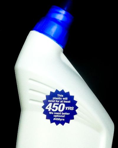 Close-up photograph of a white plastic bottle, shaped like a toilet cleaner container. On the front of the bottle is a blue, sunburst-shaped sticker that says, 'This plastic will exist for at least 450 YRS. We need better options! #450yrs.'