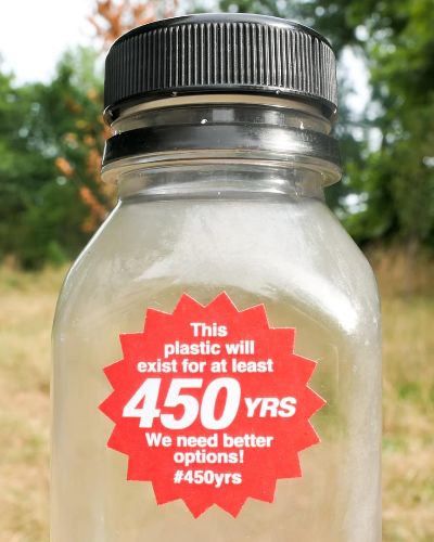 A plastic bottle with a red, starburst-shaped sticker on its front that reads: 'This plastic will exist for at least 450 YRS. We need better options! #450yrs. In the background is a blurry green field.'