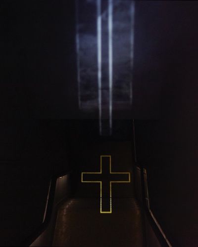 Photograph taken from atop a dark stairwell showing the outline of a crucifix projected onto the floor at the bottom of the stairwell along with the vertically oriented beams of light, visible in midair, that constitute it.
