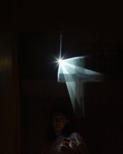 Photograph of a barely lit room with a person taking a selfie at the bottom-middle of the frame and a clearly defined crucifix seemingly floating above her left shoulder. The floating crucifix emanates from a singular point of bright light in the middle of the frame.