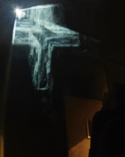 Photograph; it is extremely dark, on the right is an inkling of an illuminated handrail indicating a staircase. In the middle is a floating, cloudy, illuminated crucifix in three dimensions; towards the bottom right, if one squints, one can make out a viewer in the darkness looking up at the ghostly crucifix