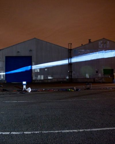 Nighttime photograph showing a large warehouse in the background onto which a large oval of white rectilinear geometric forms is projected. In the foreground is a large, empty parking lot with projection equipment strewn about.