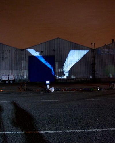 Nighttime photograph showing three large warehouses in the background onto which is projected a few dozen white butterfly-like forms, one behind the other in a figure-eight-ish configuration. The figure-eight-ish configuration is not flat but rather rotated along the y-axis in 3D space. In the foreground is a large, empty parking lot with projection equipment strewn about, and the sky behind the warehouses is a dark shade of burnt orange.