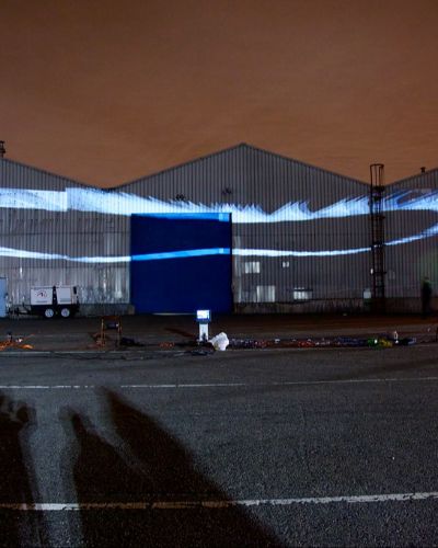 Nighttime photograph showing three large warehouses in the background onto which is projected several dozen white oval-esque forms, one behind the other in a flying-V formation. The flying-V formation is not flat but rather angled downward. In the foreground is a large, empty parking lot with projection equipment strewn about, and the sky behind the warehouses is a dark shade of burnt orange