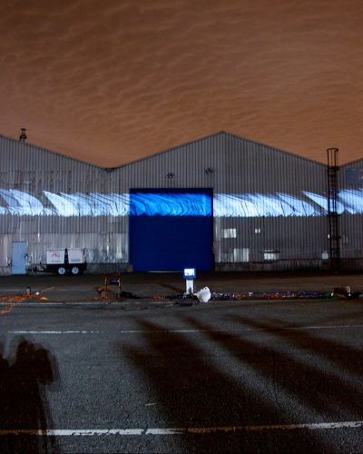 Nighttime photograph showing three large warehouses in the background onto which is projected a large white, swoosh-like form. In the foreground is a large, empty parking lot with projection equipment strewn about, and the sky behind the warehouses is a dark shade of burnt orange.