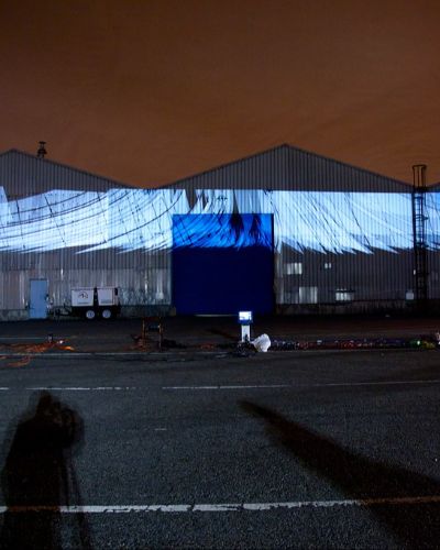 Nighttime photograph showing three large warehouses in the background onto which is projected over 100 white sail-shaped forms, one behind the other along a cinched oval-shaped path. In the foreground is a large, empty parking lot with projection equipment strewn about, and the sky behind the warehouses is a dark shade of burnt orange.