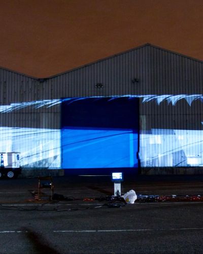Nighttime photograph of three large warehouses, each with a triangular house-like top, with a large, 200+ foot projection of what looks like a series of graphically designed white sails following each other around a circular loop.