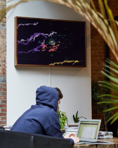 Photograph taken inside a cafe. There's a person with a blue hoodie and a computer at a wooden table in the foreground and an Enter The Machine artwork hanging in the background.