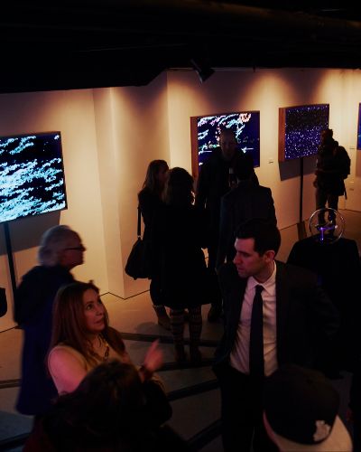 Photograph of a gallery opening taken from above showing roughly 20 people looking at illuminated artworks hanging on a wall; the gallery space is dimly lit.