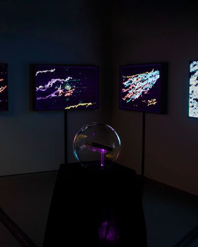 Photograph of a dimly lit, empty gallery space with four illuminated artworks hanging on the wall in the background and a glass globe on a dark pedestal in the foreground.