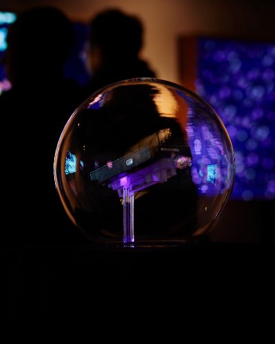 Photograph of a hard drive seemingly floating inside a glass sphere, reflecting the light of the illuminated paintings around it; in the background are two blurry figures looking at artwork.