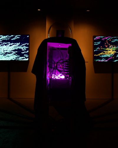 Photograph in a dark art gallery, in the center of which is an open cavity of a box showing electrical wires going hither and thither, backlit by a purple light and shrouded by black cloth on the sides. On top of the box is a glass orb and behind it, flanked on both sides are illuminated artworks hanging on barely lit walls.
