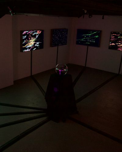 Photograph of a dimly lit, empty gallery space showing five illuminated works hanging on a wall. In the middle of the space is a glass globe on a black pedestal, from the base of which run thick black wire molding that connects to the base of each illuminated artwork.
