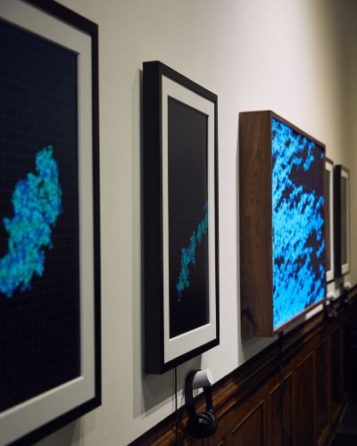 Close-up photograph of three teal blue artworks hung on a white wall, taken at a 45-degree angle such that the black metal frames are prominently shown.