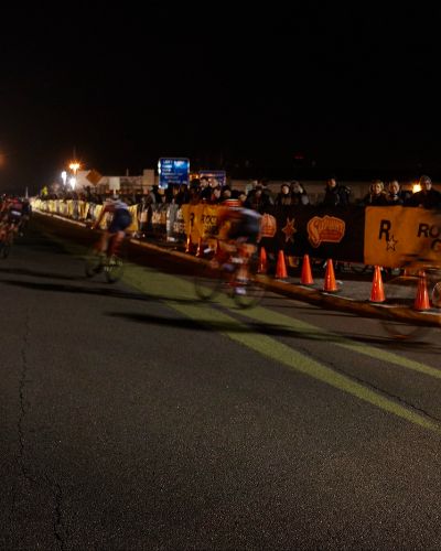 Nighttime photograph of a bike race; several riders are seen rounding a corner and riding over a large, projected yellow arrow.