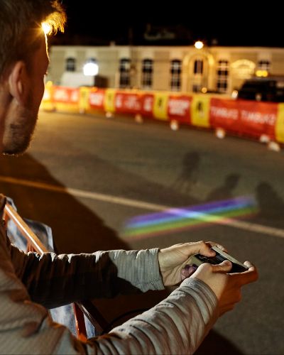 Nighttime photograph of a man, on the left side of the frame, with an (original) Nintendo controller in his hands. On the right side, projected onto the race course behind him, is a multicolored, striped rectangle.