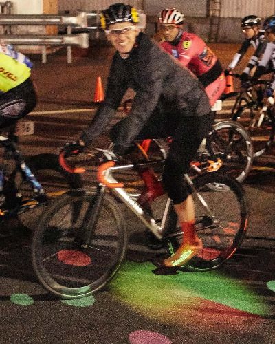 Nighttime photograph of a cyclist, in a race, looking straight at the camera (smiling), as he and his bike pass over an illuminated projection of a circular cornucopia of multicolored shapes that are projected on the race track. He is one of a bunch of cyclists passing over this colorful projection.