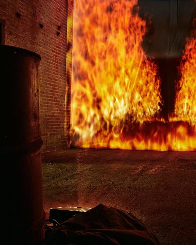 Close-up nighttime photograph of an oil drum on the left and a (projected) raging firescape in the background towards the right