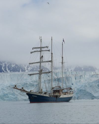 Photograph of an arctic landscape: cold gray sky, snow-covered mountains in the far background, a 20-30 meter high glacier in the near-background, and a old-timey sail ship anchored in calm waters on the left of the frame.