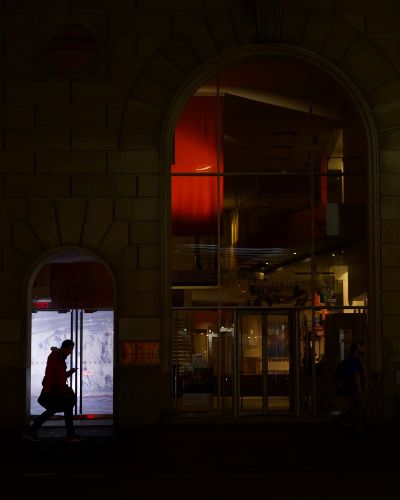 Very dark nighttime photograph of a silhouetted figure walking down the street looking at their phone. Behind him is a projected image of glaciers in a small storefront window.