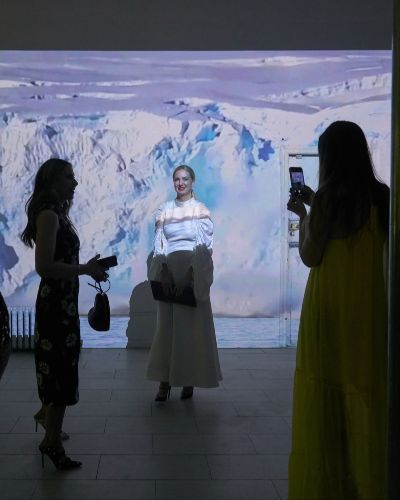 Photograph of four people in a dark gallery space. Only one is illuminated, who appears to be a blond-haired woman, very nicely done-up, and who is having her picture taken by someone else in the frame. She is illuminated by a projection of a glacier collapsing behind her. She is smiling.