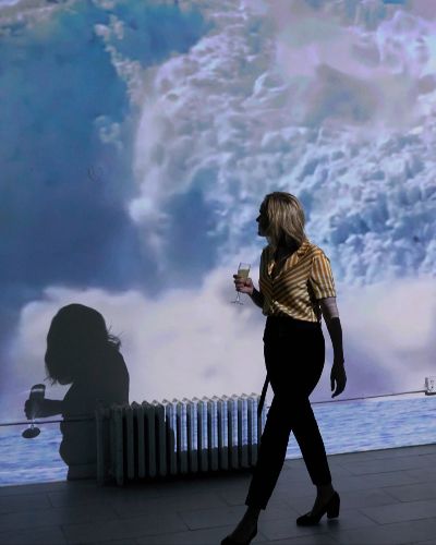 Slighly askew photograph of a women in cocktail attire, holding a cocktail, walking in front of a projection of a glacier collapsing