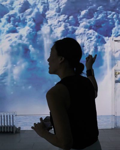 Close-up photograph of a person's head, in 3/4 profile in the middle of the frame. She is pointing to the wall behind her, onto which is projected an image of a glacier collapsing. She appears to be saying something.