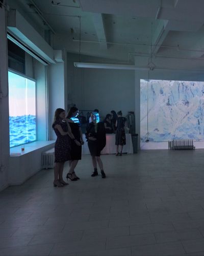 Wide shot of the inside of a gallery showing two different projected works. On the left, an image of a body of water is projected into the gallery windows. Straight back, an image of an Arctic scene&mdash;close up of a glacier&mdash;is projected. There are about a dozen people in cocktail attire milling about. On the right, a two sharply bright lights are emanating from the lens of large projectors.